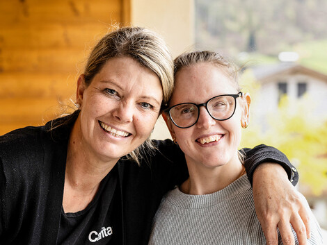 Eine Caritas Mitarbeiterin und eine Klientin mit Behinderung stehen Arm in Arm für ein Portrait zusammen.