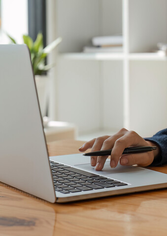 Eine Frau sitzt an einem Holztisch und hat ein Buch vor sich aufgeschlagen sowie einen Laptop aufgeklappt.