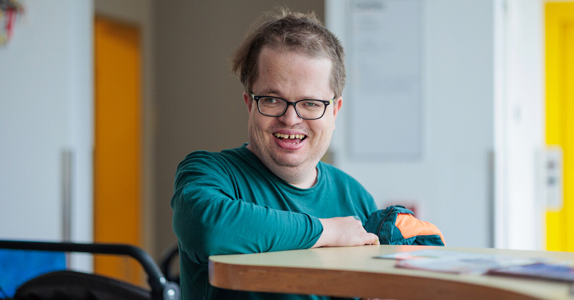 © Ruben Lackner Ein lachender Mann mit Behinderung und Brille steht in einer Tagesstätte für Menschen mit Behinderung am Empfangstresen.