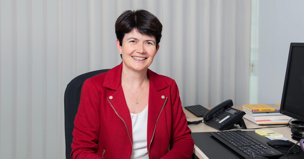 Portrait einer lachenden Frau, die an einem Büroschreibtisch sitzt und eine rote Jacke trägt.