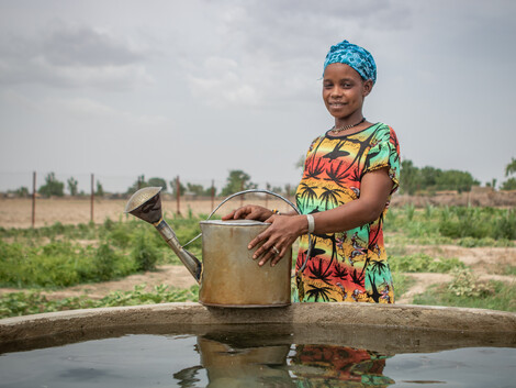 Portrait einer jungen Frau in traditioneller afrikanischer Kleidung die eine Blechgieskanne auf einem Bewässerungsbrunnen abgestellt hat. Im Hintergrund ist ein Gemüsefeld zu sehen.