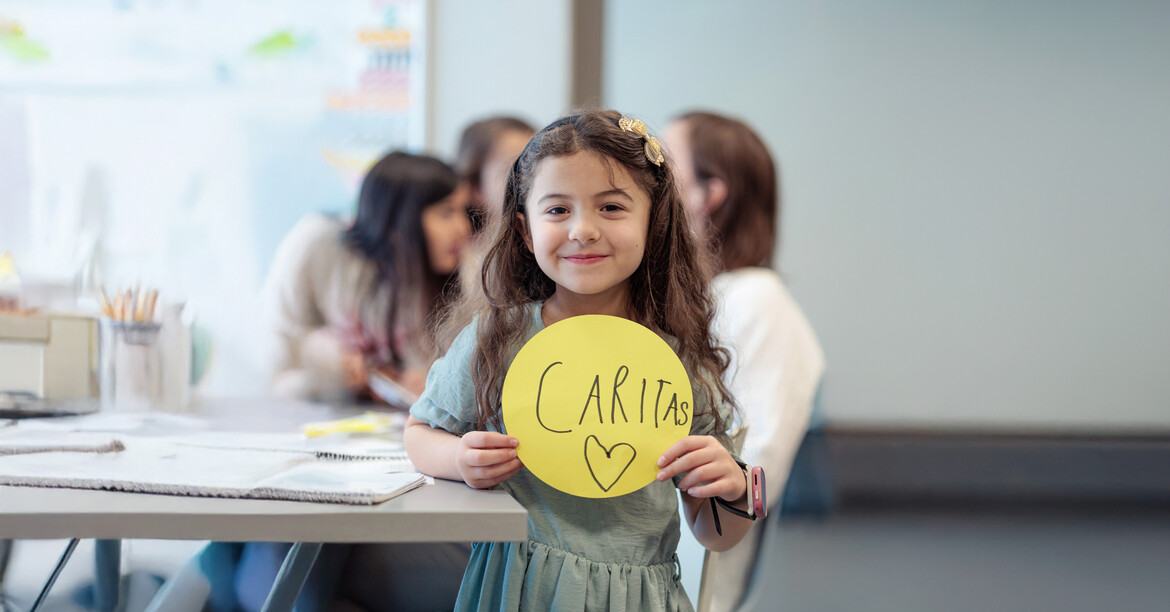 Ganzkörper Portrait eines Mädchens in einer Lernbetreuungseinrichtung. Sie präsentiert ein rundes, gelbes Papier auf das sie "Caritas" geschrieben und ein Herz gemalt hat.
