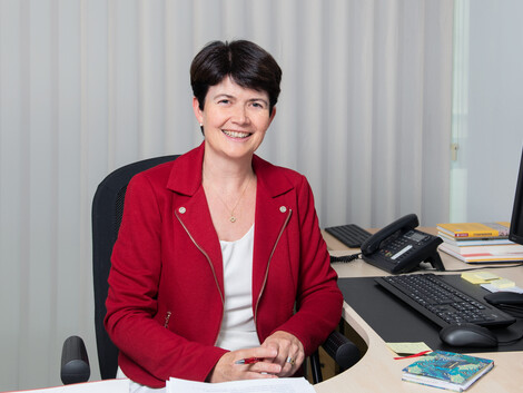 Portrait einer Frau mit roter Jacke an einem Schreibtisch sitzend.