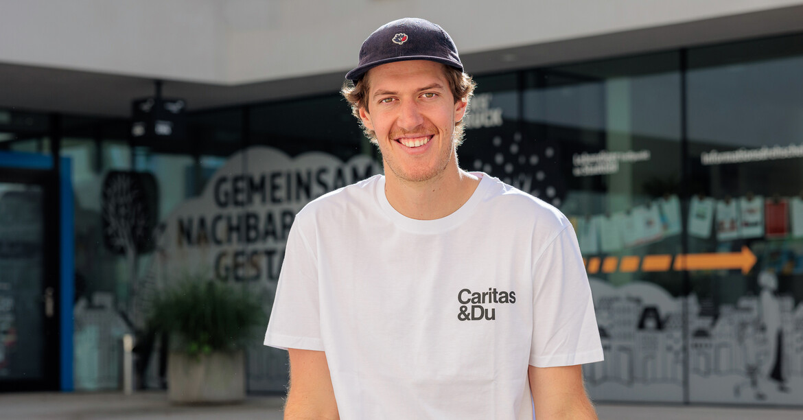 Einer junger Mann mit Shildkappe und weißem Caritas T-Shirt sitzt auf einer Bank vor einem Gebäude.