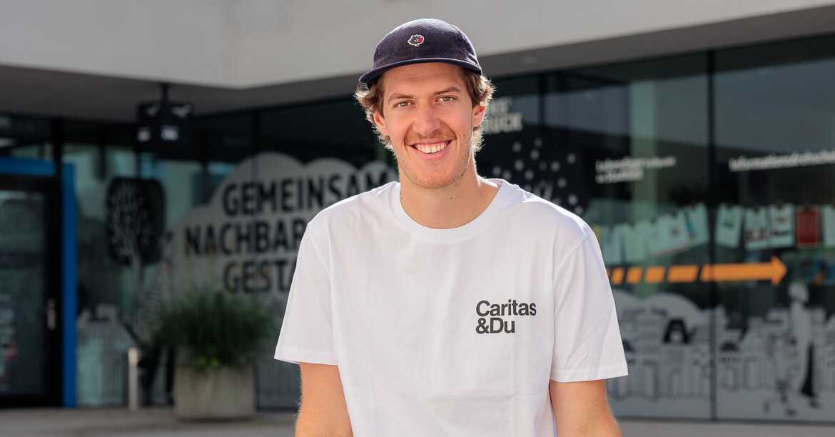 © Ruben Lackner Einer junger Mann mit Shildkappe und weißem Caritas T-Shirt sitzt auf einer Bank vor einem Gebäude.