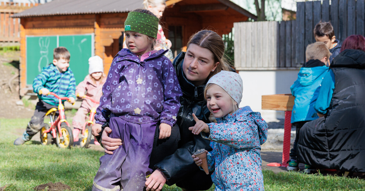 In einem Garten geht eine Kindergärtnerin in die Hocke, um mit zwei Kindern auf Augenhöhe zu sein.